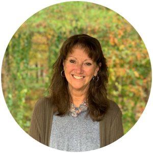 Photo of PJC's CEO, Pam Newcombe, standing in front of trees wearing a grey shirt
